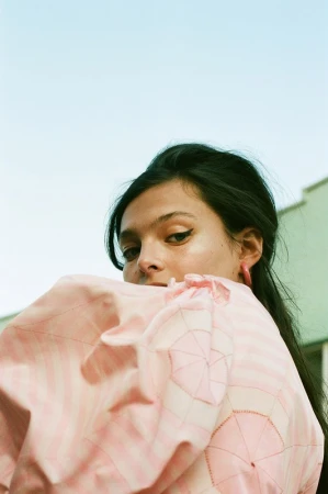 Close-up portrait of a brunette woman in a pink dress with umbrella pattern against a light blue and green exterior background.