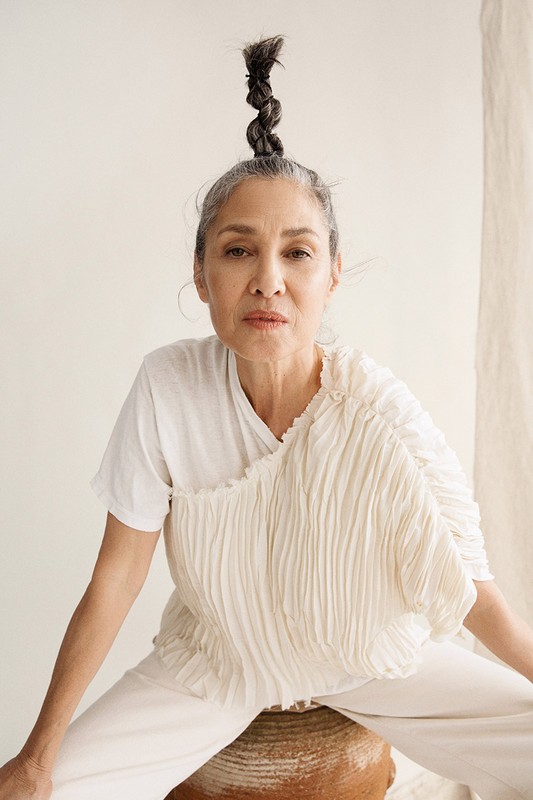 Portrait of an older adult female with braided hair posing in white clothing seated on a brown vase against an off-white background.