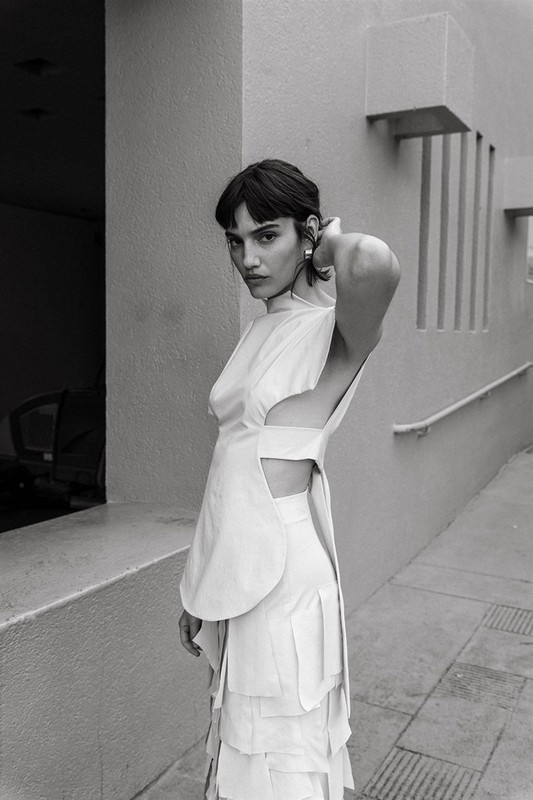 Monochromatic portrait of a woman in avant-garde layered dress with structural elements, minimalist outdoor setting, soft light, and striking pose.