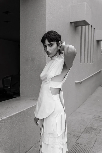 Monochromatic portrait of a woman in avant-garde layered dress with structural elements, minimalist outdoor setting, soft light, and striking pose.