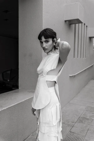 Monochromatic portrait of a woman in avant-garde layered dress with structural elements, minimalist outdoor setting, soft light, and striking pose.