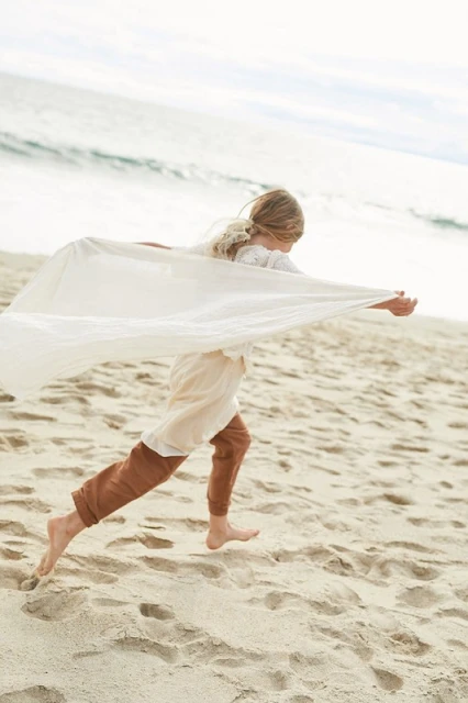 Young girl with blond hair running barefoot on sandy beach, holding out a white sheet, with ocean waves in the background.