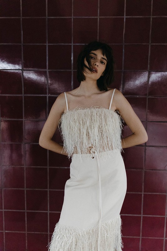 A woman in a white fringed top and matching skirt poses against a tiled wall, showcasing fashion and style with a neutral color palette.