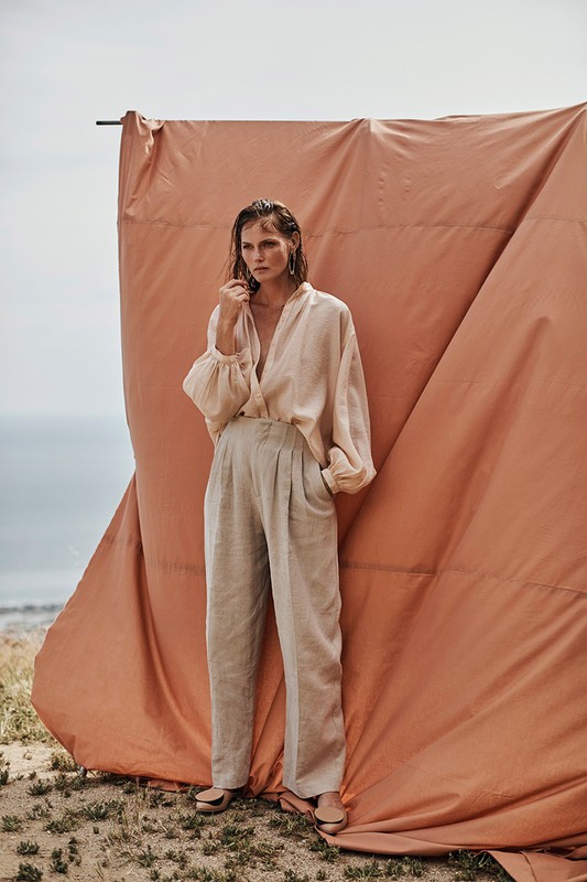 A fashionable woman poses in a linen outfit against a draped peach backdrop near the ocean, showcasing neutral, earthy tones.