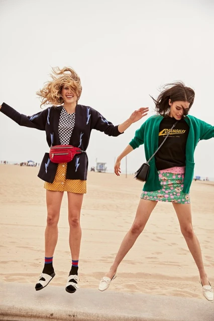 Two women jumping on a beach wearing mini skirts and cardigans. They are smiling, with one woman blonde and the other brunette.