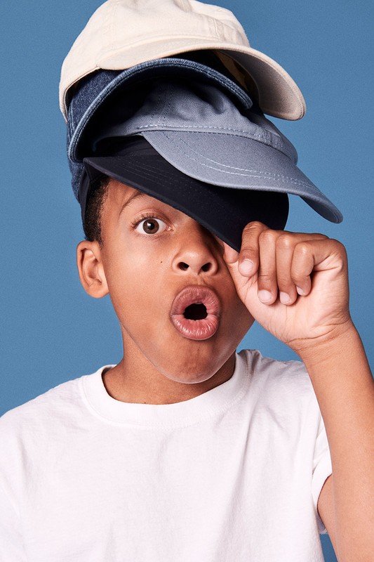 Young boy balancing four baseball caps on his head with a surprised facial expression against a blue background in a studio shot.
