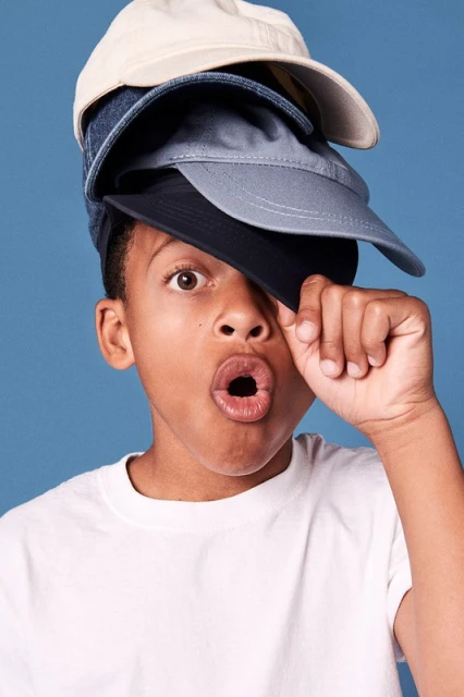 Young boy balancing four baseball caps on his head with a surprised facial expression against a blue background in a studio shot.