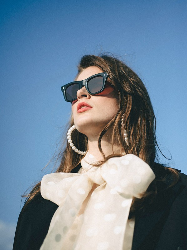 A stylish woman with dark sunglasses and pearl earrings poses in front of a clear blue sky, wearing a black jacket and polka-dot bow blouse.