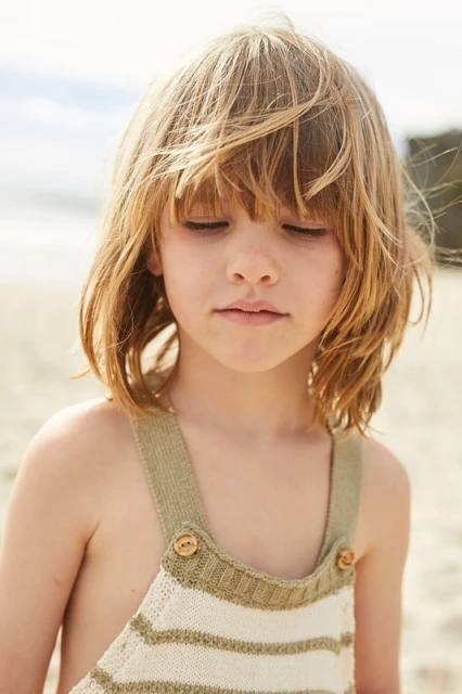 Close up of child with blonde hair in striped overalls, looking down on a sandy beach with the sky visible in the background.