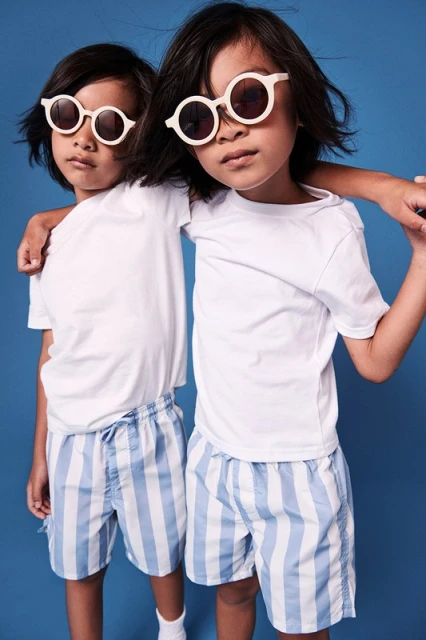 Two young children in matching white t-shirts and blue striped shorts pose in front of a solid blue studio backdrop, wearing round sunglasses.