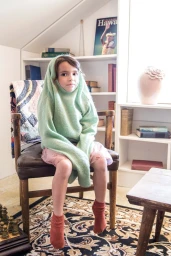 Young child seated in a wooden chair wearing an oversized mint green sweater pulled over head