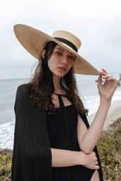 Woman in wide-brimmed straw hat and black ribbed top standing outdoors by the ocean with a rocky coastline