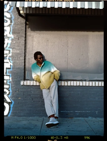 A Black man leaning against a brick storefront wearing a green gradient cardigan and light trousers, styled by Steph Ashmore.