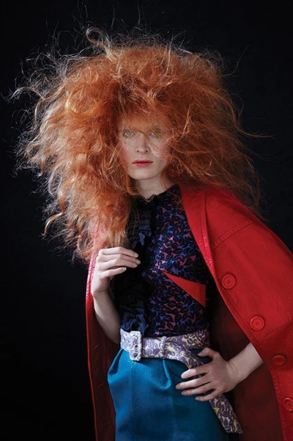 Fashion shot of a red-haired model wearing a colorful top, blue skirt, and red coat against a dark backdrop in a photo studio.