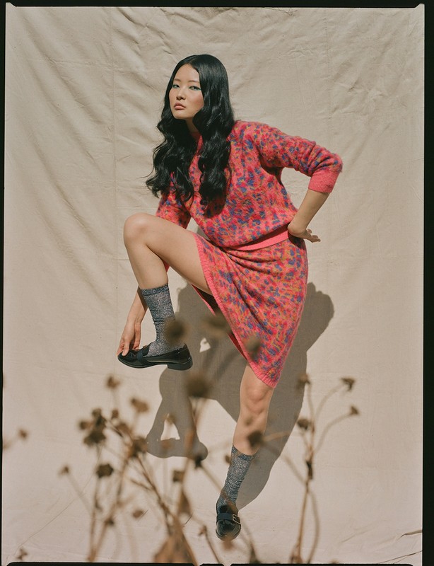An Asian woman poses in a matching pink patterned sweater and skirt, knee raised, wearing gray socks and black shoes against neutral backdrop.