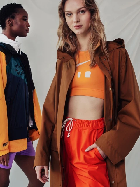 Two models pose indoors wearing Nike branded sportswear in a studio setting with a white backdrop and bright lighting, facing forward.