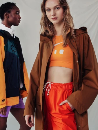 Two models pose indoors wearing Nike branded sportswear in a studio setting with a white backdrop and bright lighting, facing forward.