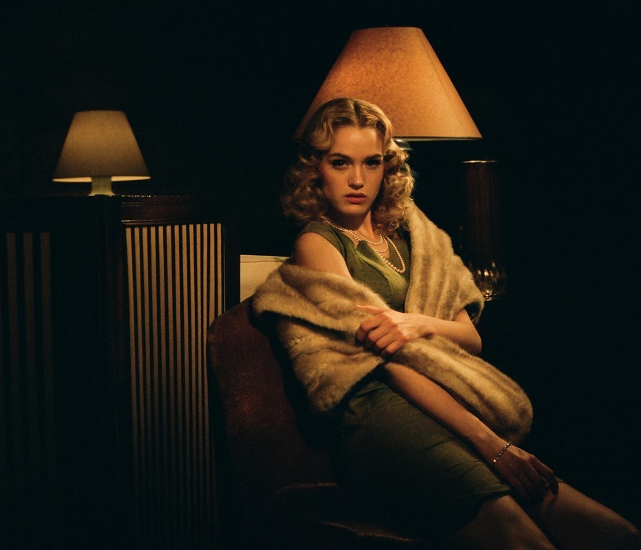 A woman with blonde curly hair, wearing a green dress and faux fur stole, poses indoors under the warm glow of lamps.