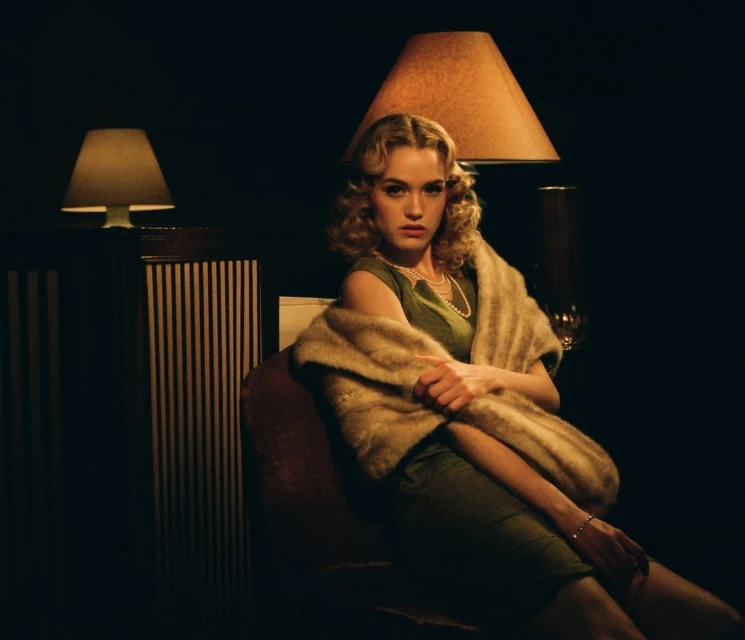 A woman with blonde curly hair, wearing a green dress and faux fur stole, poses indoors under the warm glow of lamps.