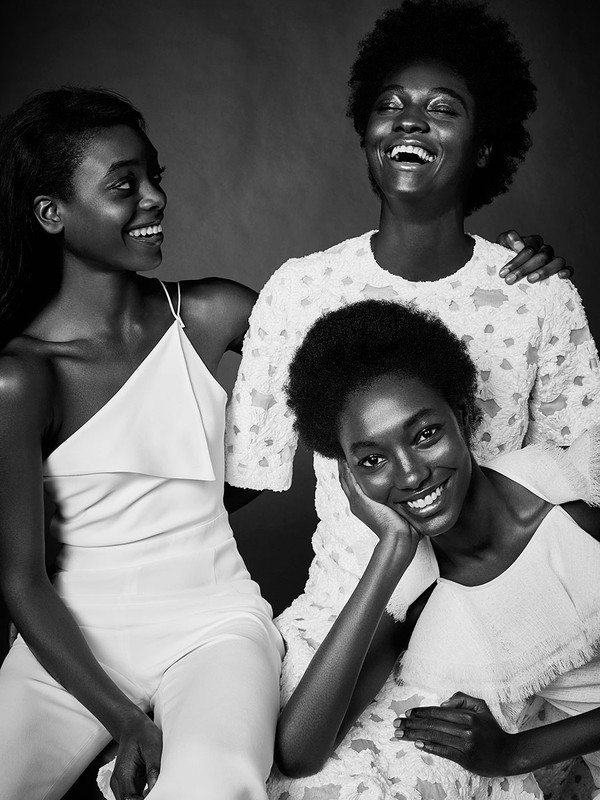 Black and white portrait of three smiling Black women in white outfits posing together in a studio setting against a dark background.