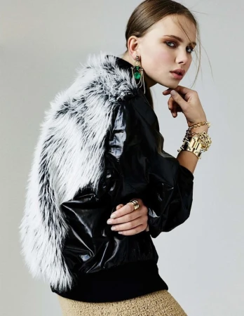Model in black jacket with white fur back panel posing against a plain grey background with gold jewelry