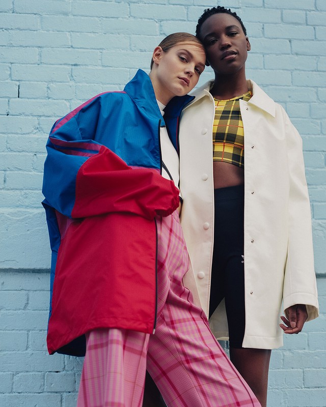 Two women pose in front of a blue brick wall, wearing colorful jackets and pants, with one woman resting her head on the other's head.