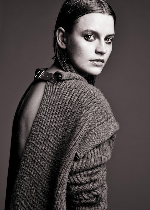 Portrait of a young woman with wet-look hair wearing a dark sweater, gazing over her shoulder in a studio setting.