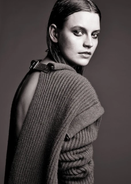 Portrait of a young woman with wet-look hair wearing a dark sweater, gazing over her shoulder in a studio setting.