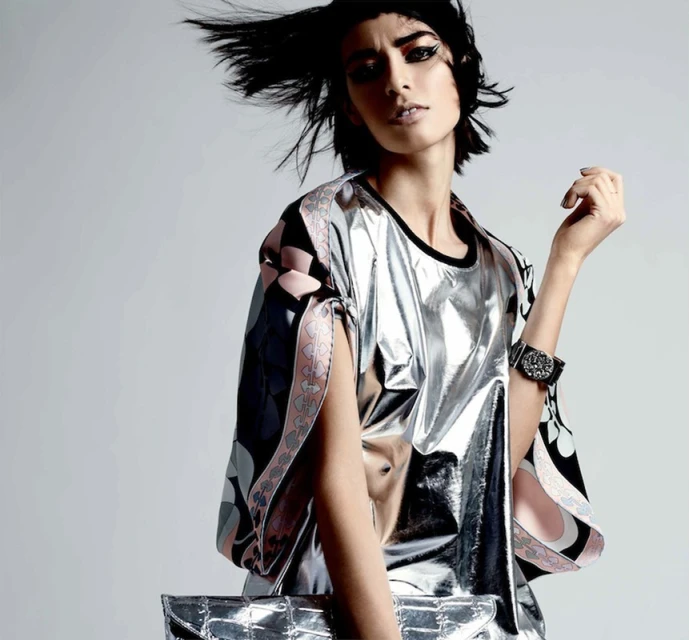 Fashion portrait of a female model wearing a shiny silver blouse, holding a silver clutch, with dark hair and eye makeup in a photo studio.