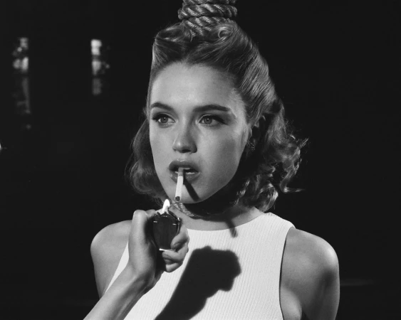 Monochrome portrait of a fair-skinned woman with retro hairstyle lighting a cigarette, with rope above her head, and hand holding lighter.