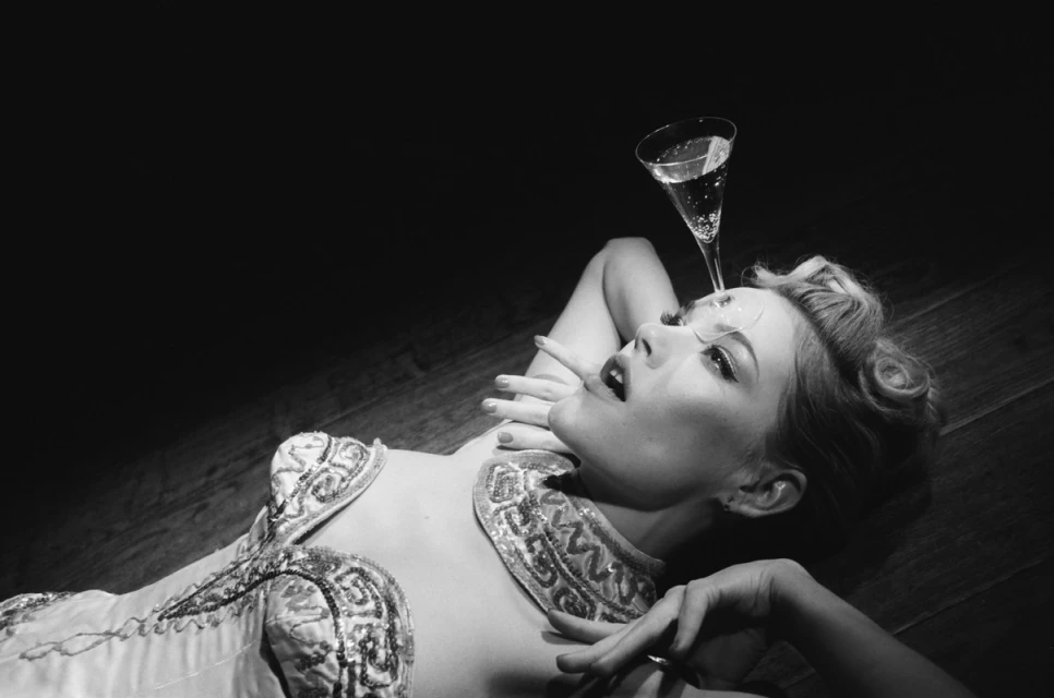 Black and white portrait of a woman lying on her back with a champagne glass balanced on her forehead in an indoor setting.