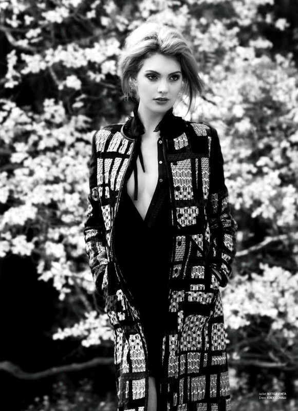 Black and white portrait of a fair-skinned woman with blonde hair, wearing a patterned jacket and dress, posing outdoors against a floral background.