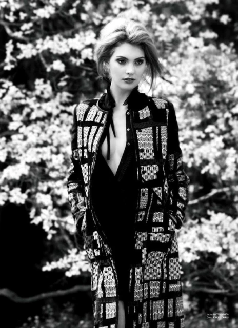 Black and white portrait of a fair-skinned woman with blonde hair, wearing a patterned jacket and dress, posing outdoors against a floral background.