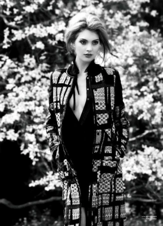 Black and white portrait of a fair-skinned woman with blonde hair, wearing a patterned jacket and dress, posing outdoors against a floral background.