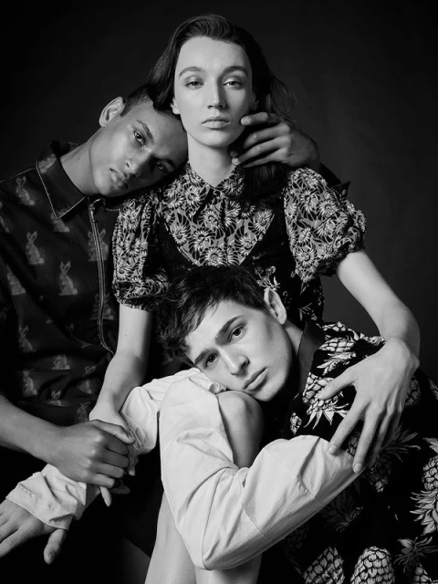 Black and white portrait of three people wearing patterned shirts in a close studio shot with a dark background and diffused lighting.