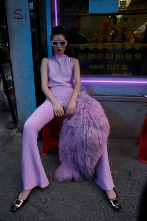 Stylish Asian model in pink pantsuit and white sunglasses sitting in front of a vintage hair salon in an urban setting.