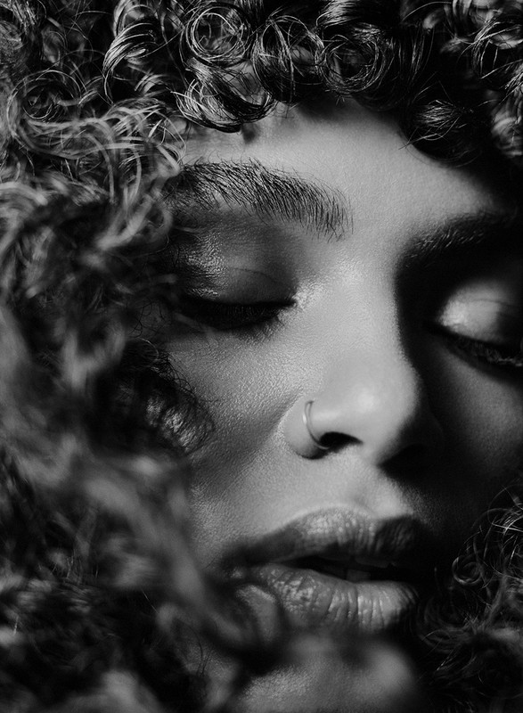Striking black and white close-up portrait of a woman with curly hair and closed eyes, featuring a nose ring and subtle facial lighting.