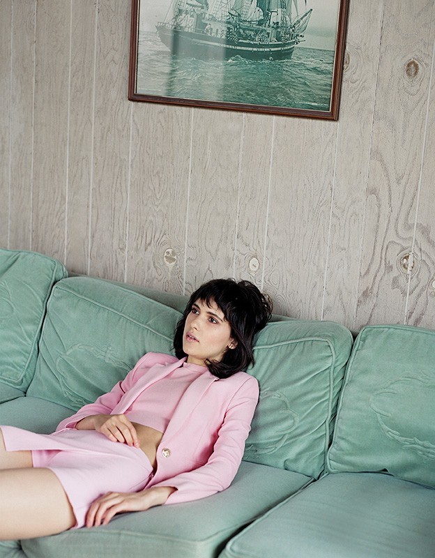 Brunette woman in a pink suit and crop top, lying on a plush green couch with a framed nautical print hanging on the wall.
