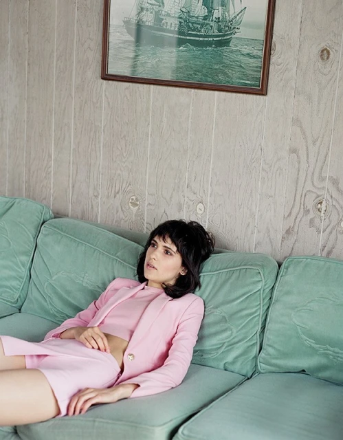 Brunette woman in a pink suit and crop top, lying on a plush green couch with a framed nautical print hanging on the wall.