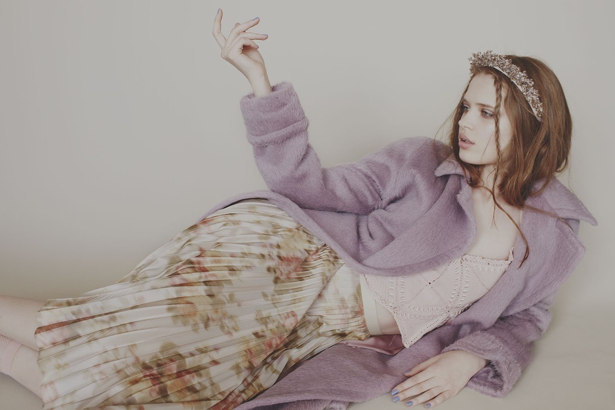 Young woman in lilac coat and floral skirt reclines in a studio. She wears a beaded headband and her hand is raised in a pose.