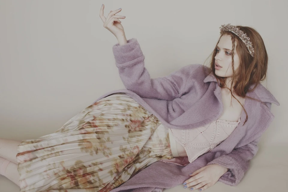 Young woman in lilac coat and floral skirt reclines in a studio. She wears a beaded headband and her hand is raised in a pose.
