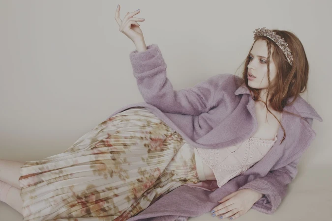 Young woman in lilac coat and floral skirt reclines in a studio. She wears a beaded headband and her hand is raised in a pose.