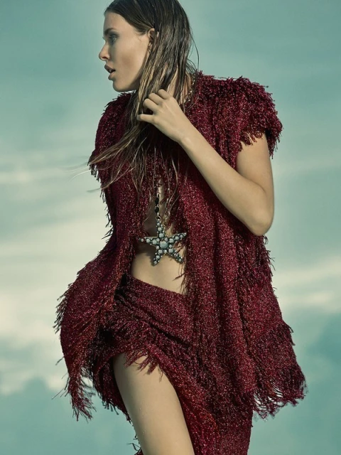Profile shot of a female fashion model with wet hair wearing a red, fringed outfit with a star-shaped necklace against a sky background.