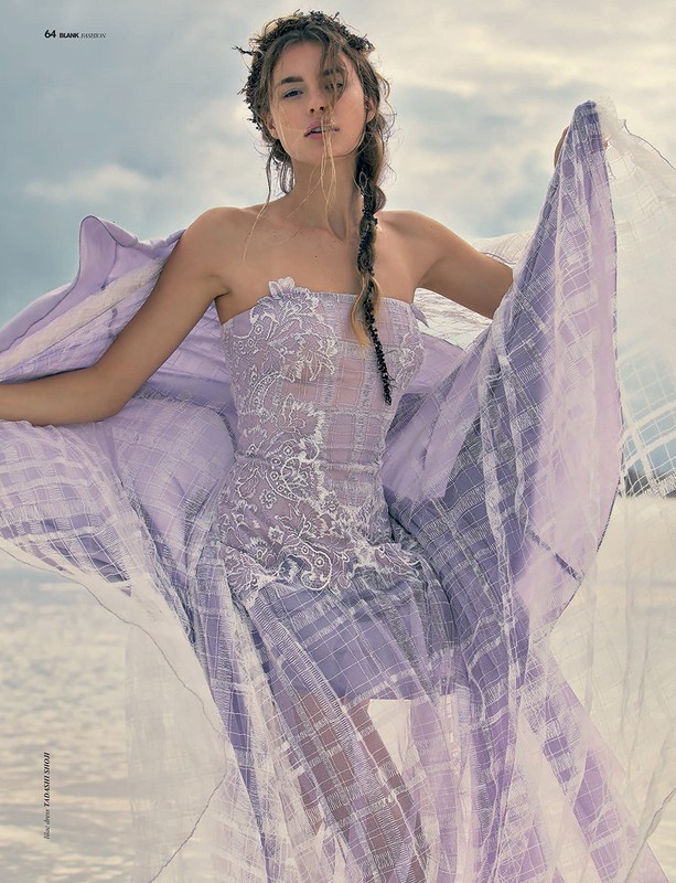 Model in a lavender strapless dress with sheer fabric billowing in the wind against an overcast sky and water backdrop