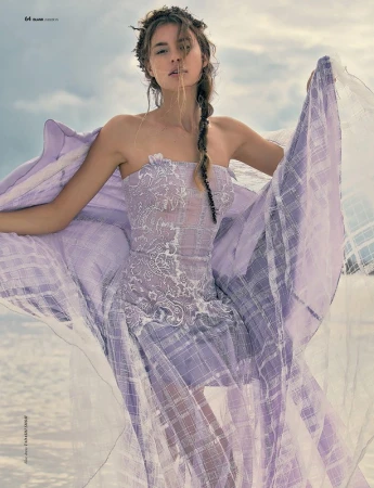 Model in a lavender strapless dress with sheer fabric billowing in the wind against an overcast sky and water backdrop