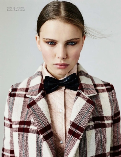 Close-up of a young adult woman wearing a plaid coat and bow tie looking at the camera in a studio environment with neutral background.