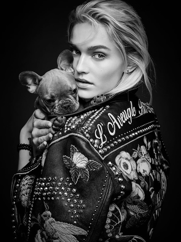 Blonde woman in black jacket holds a French bulldog puppy in a black and white portrait against a dark background.