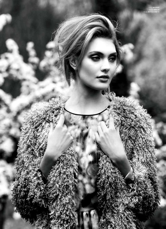 Black and white portrait of a fashionable woman with blonde hair wearing a textured coat against a blurred outdoor backdrop with foliage.