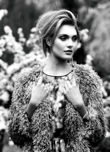 Black and white portrait of a fashionable woman with blonde hair wearing a textured coat against a blurred outdoor backdrop with foliage.