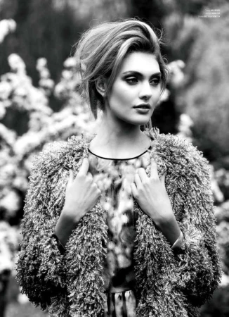 Black and white portrait of a fashionable woman with blonde hair wearing a textured coat against a blurred outdoor backdrop with foliage.